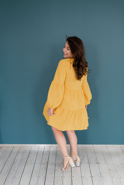 Bee, founder and owner of BusyBSocial, her body faces away from the camera and she smiles with her eyes closed looking back wearing a yellow dress in front of a blue background