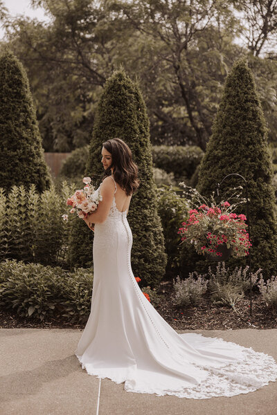 A bride in a sleeveless gown with a train, holding a bouquet, stands amidst a garden.