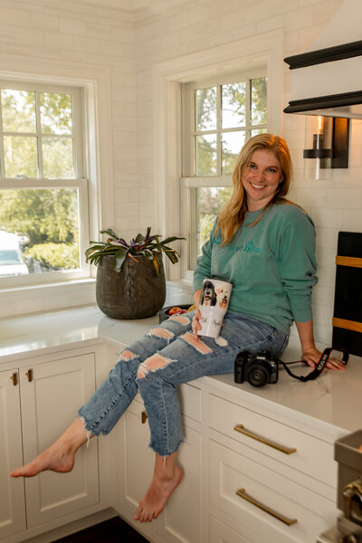 Image of Brook sitting on a kitchen counter top, smiling at the camera while holding travel thermos with dogs painted on it