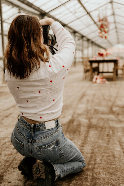 Laura, photographe de mariage en Vendée, accroupie, prend une photo de dos sous une immense verrière avec une table décorée pour un mariage au centre.