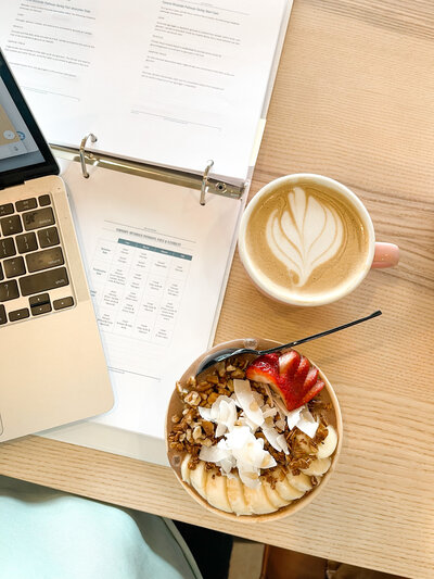 Image from above of a laptop and a binder of papers next to a smoothie bowl and a mug of coffee