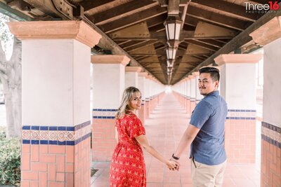 Engaged couple look back at the photographer while walking through the Santa Ana Train Station while holding hands