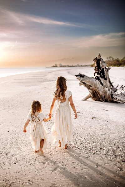 Family Photographer, two young sisters hold hands as the walk in the beach sand