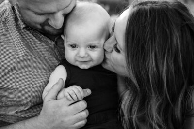 A black and white close-up of a baby being held by both parents. The baby is in the center, looking directly at the camera, while the mother kisses the baby’s cheek and the father smiles down at the baby.