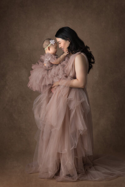 Mother in a flowing blush gown lovingly holding her baby girl in a matching tulle dress during a studio session. Captured by a Dallas Baby Milestone Photographer specializing in motherhood portraits.