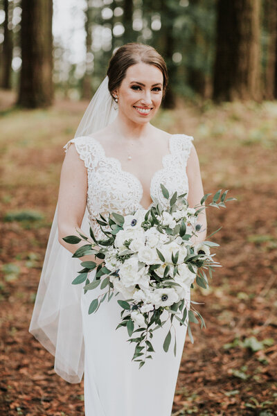 real bride wearing a custom bridal veil and holding a cascading bouquet of white flowers and greenery while standing in a forest setting.