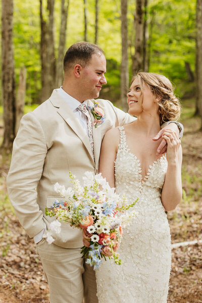 In a Hudson Valley forest, groom in tan suit embraces bride in unique lace dress with whimsical white, blue, and peach bouquet as they look into each others' eyes.