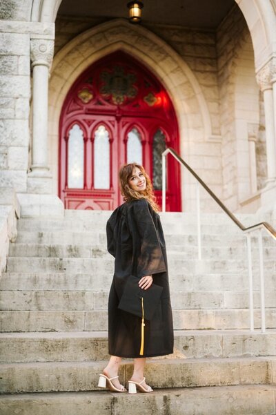 A graduate in a black gown holds a cap on stone steps at St. Edward's University smiling back. 