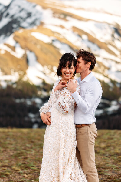 Shannon Lee, a Colorado elopement photographer, captured a joyful couple embracing in front of snowy mountains. The woman wears a lacy dress, while the man is dressed in a light shirt and pants, evoking a sense of happiness, romance, and the beauty of their intimate elopement.