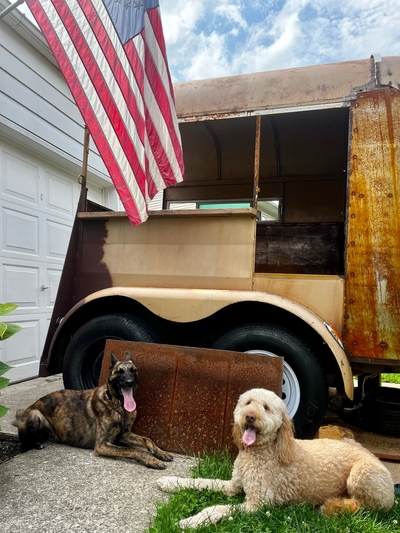 Unfinished mobile bar in progress during conversion stage. Two dogs sit out front next to the trailer and rusty metal sheets. An American flag is in the top corner.