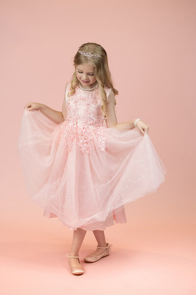 Young girl in a delicate pink dress with lace details, holding the skirt out. She wears a tiara and smiles softly against a matching pink background.