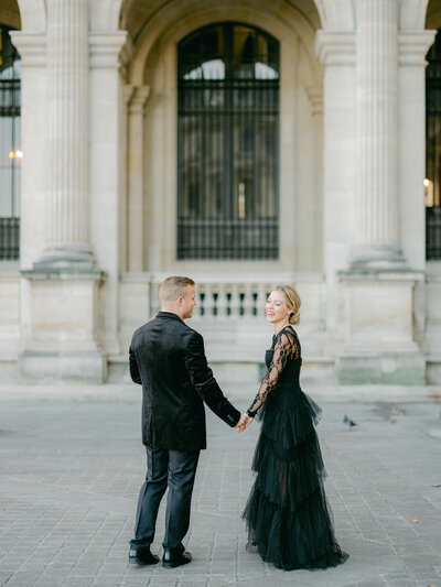 a woman and man in a black suit and tulle dress looking at each other and smiling while standing in front of the louvre