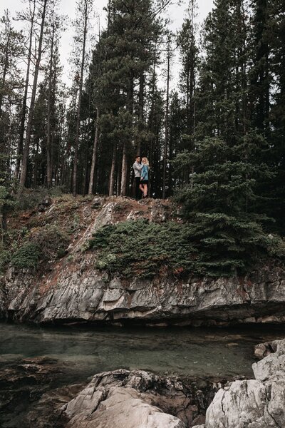 young couple stands on the edge of a creek cliff in Kananaskis, facing each other while the man puts his hand on the woman's cheek