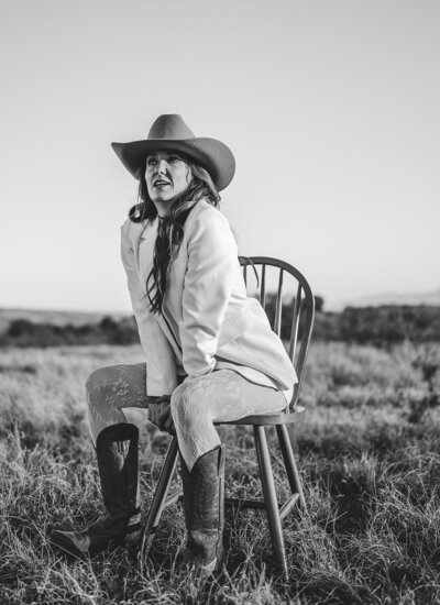 Black and white photo of Juliet Trnka, outdoors, sitting on a chair, wearing a cowboy hat and boots and a beige blazer-part of the Direct Knowing photoshoot