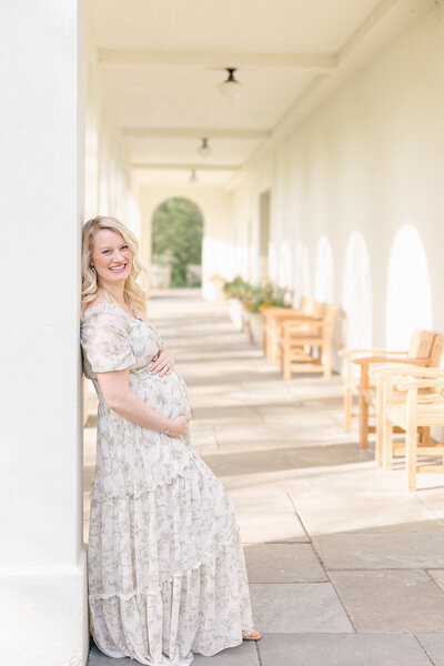 Pregnant mother in green floral dress leans against post at Newfield's Lilly House during session with Indianapolis maternity portrait photographer