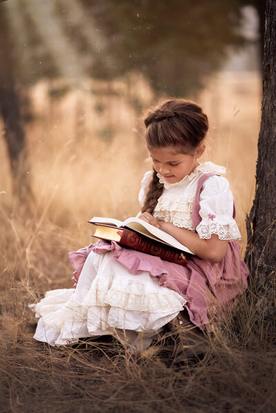 Beautiful portrait of a little girl with long braided hair wearing a vintage dress crouched against a tree reading a book. Captured by Brittany Danielle Photography.