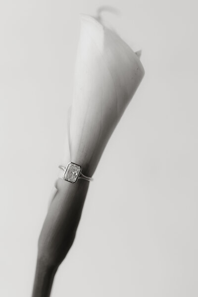Close-up of an engagement ring resting on a calla lily, photographed by Rainstorm Photo & Video in soft black and white tones to highlight its elegant, minimal detail.