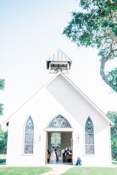 Exterior photo of a white church with stained glass windows and a couple getting married through the doorway