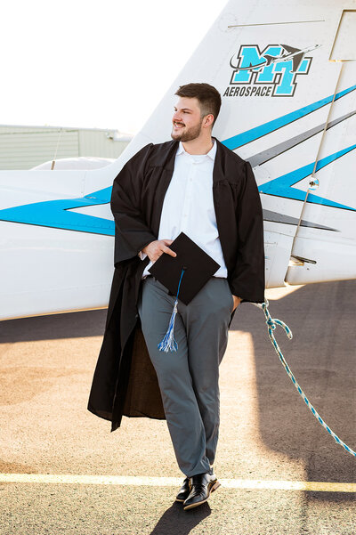 Senior guy in suit wearing graduation down and golding cap standing beside a plane on the aviation field for MTSU