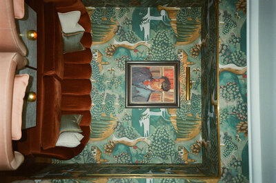 Red couch in front of illustrative green wallpaper with a painting of a portrait in the middle