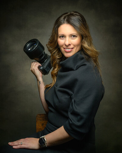 This professional portrait features Ana-Maria Manoila, an international award-winning pet photographer based in Vancouver. Ana-Maria is elegantly dressed in black, seated confidently while holding her camera. Her warm smile and poised demeanor reflect her passion and expertise in pet photography. The dark, textured backdrop adds a sophisticated touch, highlighting Ana-Maria's dedication to capturing the unique bond between pets and their owners. As the talented artist behind Pets through the Lens Photography, Ana-Maria’s skill and artistry are evident, showcasing her as a leading figure in the world of pet photography.