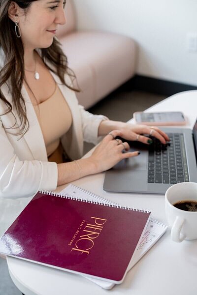 Pierce PR team member working on a laptop at a white table with a branded maroon notebook, smartphone, and coffee cup in view.