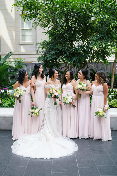 A bridal party wearing soft pink gowns smiles and laughs with their friend, the bride, while posing for bridal party portraits at their DMV wedding