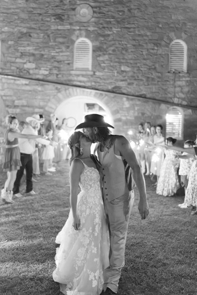 A man and woman kiss in front of a stone barn. Their friends are holding sparklers behind them. 