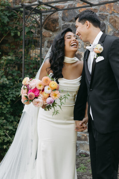 Bride laughing with her groom during outdoor portraits while holding a colorful garden-style bridal bouquet filled with dahlias, roses, and ranunculus in peach, pink, and yellow tones against a stone wall backdrop