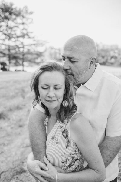Husband and wife black and white portrait with husband leaning in close, wraping his arms around wife while she smiles at the camera.
