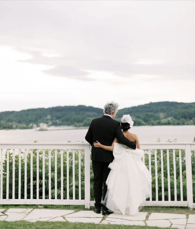 A bride and groom stand overlooking a lake.