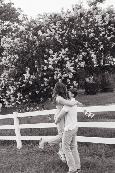 Couple embracing in front of a white fence with blooming trees in the background — black-and-white country engagement photo captured in Alabama by EC Photography