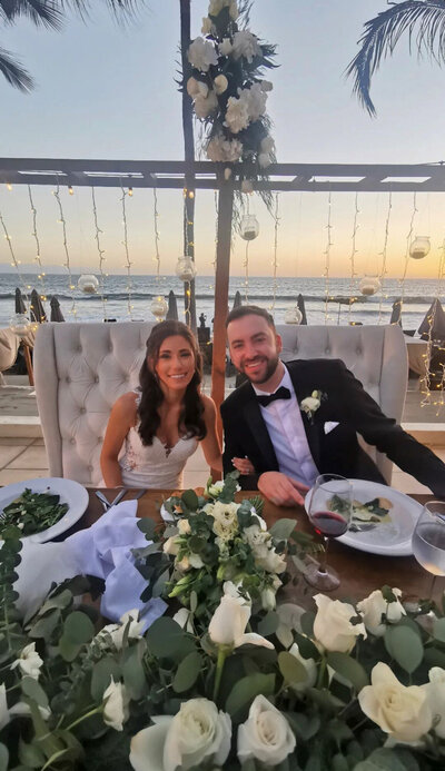 Bride and groom sitting down on their reception chair and table with a beach view behind