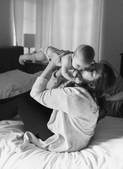 Mother sitting on bed holding baby up and kissing them at Austin family photoshoot