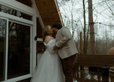 bride and groom sharing a romantic kiss on the wooden deck of a modern A-frame cabin, surrounded by tall bare trees and twinkling string lights on an overcast day. The bride wears an off-the-shoulder tulle gown with lace detailing, while the groom is dressed in a light beige blazer and dark trousers. 