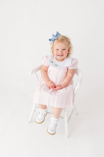 Little girl in a pink dress with a blue bow sitting on a white chair smiling at the camera