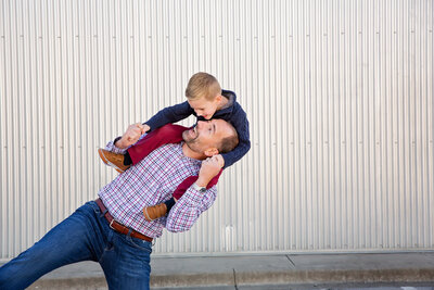 A father laughs while carrying his young son on his shoulders and leaning to the side.