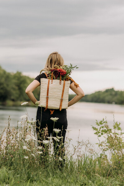 Lauren Hughes is wearing a wicker backpack with a bouquet in it. She's staring off over a river with her hands on her hips.