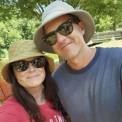 Testimonial phone photo of a husband and wife both wearing sunglasses and hats, expressing their satisfaction with Dogology University training services