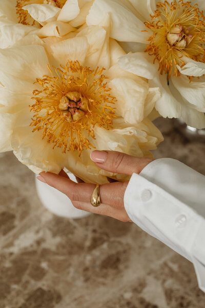 A hand gently cradles a bouquet of creamy peonies with golden centers, wearing a white shirt and gold ring.