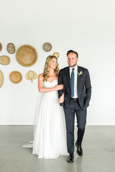 A bride and groom share a lighthearted moment inside Newport Beach House, standing against a crisp white wall decorated with woven baskets — a perfect blend of modern and coastal charm.