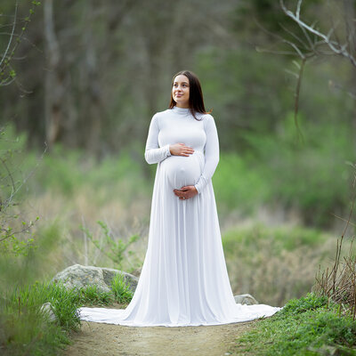 A dark haired pregnant woman is wearing a long white gown standing in the woods in the Hudson Valley NY