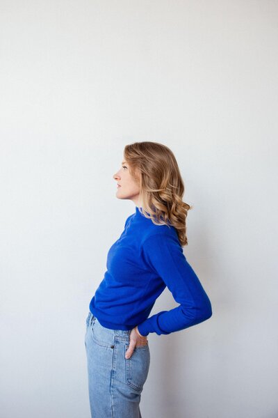 Woman in a bright blue sweater and jeans poses in profile against a clean white wall, hands resting on her hips.
