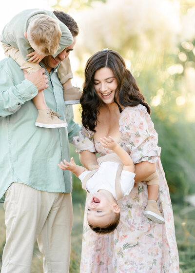 A mother with soft brown curls dressed in a peach and soft green floral gown holds her toddler son dressed in soft brown coveralls playfully upside-down while her husband holds their other toddler son on his shoulders photographed by Bay Area Family Photographer, Light Livin Photography.