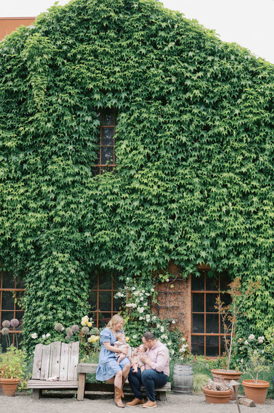 Pulled back image of family sitting on wooden benches in front of a large brick wall covered with ivy by Portland Family Photographer Emilie Phillipson Photograph 