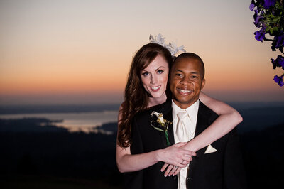 Bride hugs groom from behind in front of sunset