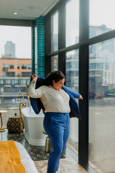 Bride wearing blue suit throws jacket on in front of window overlooking Williamsburg District