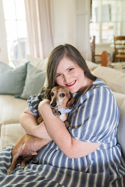 Lauren hugs her mini dachshund, Amos, in family room sofa in blue and white striped dress