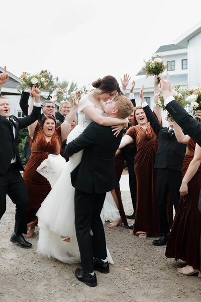 A person lifting their partner up to kiss them with their wedding parties cheer behind them 