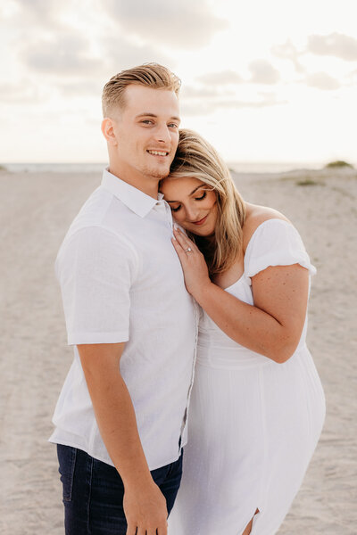 Woman wearing a white dress snuggled into his chest on the beach. Tampa area engagement photography session.